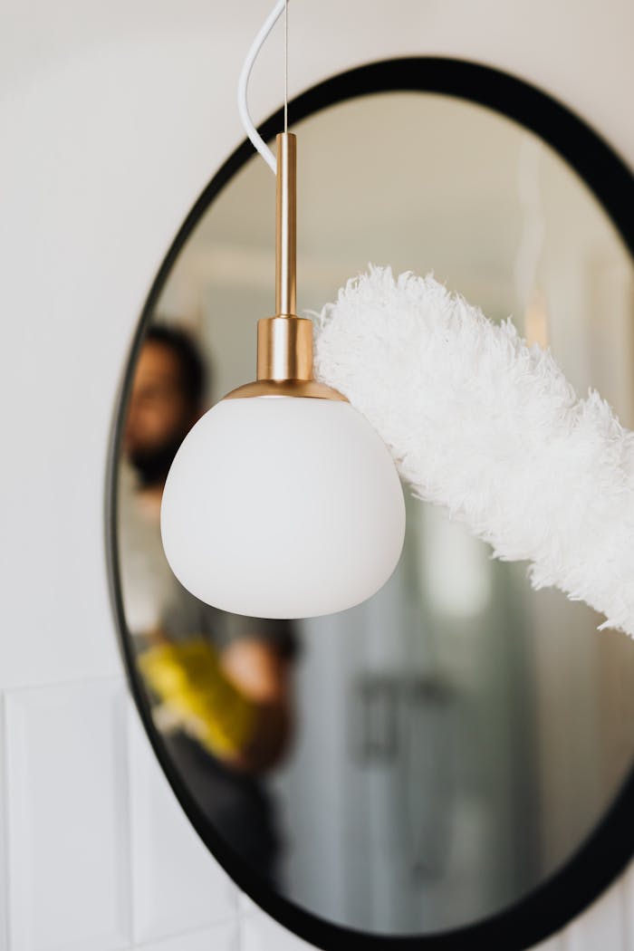 Anonymous person in casual clothes and yellow rubber glove cleaning white stylish lamp with antistatic brush dust cleaner while standing against cozy oval mirror